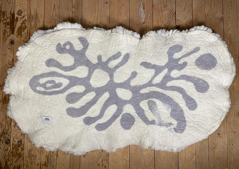 Die Liegewiese. 
Na, wenn das kein Prachtstück ist! Dieses Filzfell misst stattliche 1,75 × 1 Meter – das grösste, das je die Werkstatt der Fellfilzerei Jawoll verlassen hat.
Gefertigt aus feinster Romney-Wolle in Schafsmilchweiss, kräftig und weich zugleich, und veredelt mit einem Kreis aus Locken der Walliser Schwarznasenschafe – als wäre ein Stück Alpenpoesie eingefilzt.

Mehrere Schafe aus dem Kanton Bern haben hier ihre Wolle gespendet – und das Ergebnis ist einfach jawollig schön. Dieses Fell ist so dick, weich und robust, dass es fast schon nach draussen ruft: „Mach’s dir gemütlich!“

Ob auf dem grossen Sofa, im Bett oder direkt auf dem Boden – diese Liegewiese lädt zum Lesen, Träumen und Musiklauschen ein. Kinder lieben sie genauso wie Erwachsene, die sich zwischendurch eine sanfte Wolke unterlegen möchten.

Ein echtes Unikat – und ja, das darf man diesem Wunderwerk auch im Preis ansehen.
Mit einem Augenzwinkern: So ein Stück entsteht wohl nur einmal im Leben …